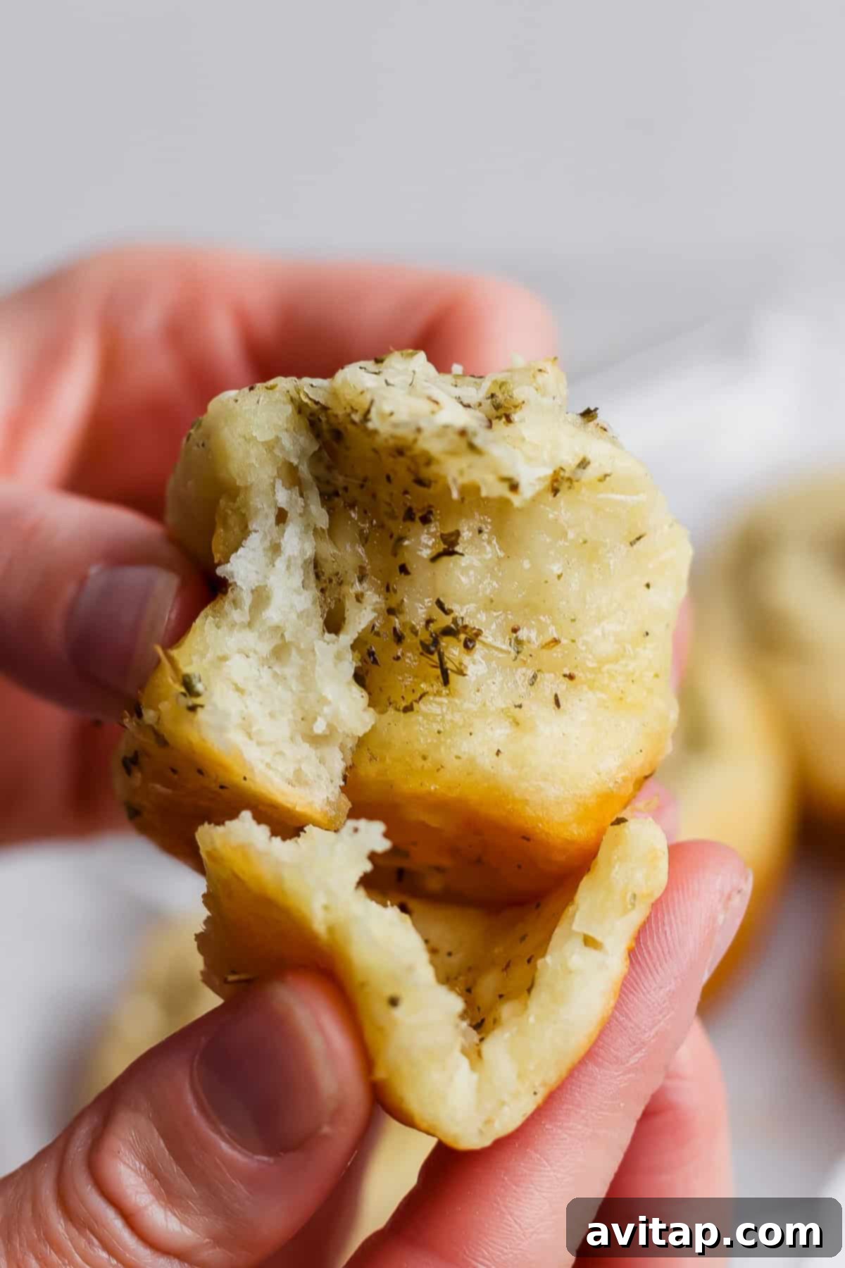A person holds and pulls apart a soft, herb-topped bread roll, revealing its fluffy interior and steam, highlighting its fresh texture.