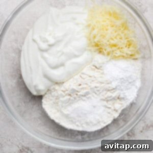 A glass bowl containing shredded cheese, plain yogurt, flour, and baking powder on a light textured surface, showing the initial ingredients for the dough.