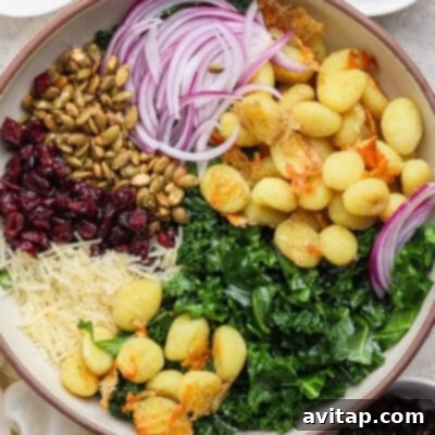 A rustic, inviting bowl filled with delightfully crispy roasted gnocchi, fresh, vibrant kale, generously shredded Parmesan cheese, tart dried cranberries, crunchy pumpkin seeds, thinly sliced red onions, and toasted breadcrumbs, all perfectly blended into a harmonious and flavorful salad.