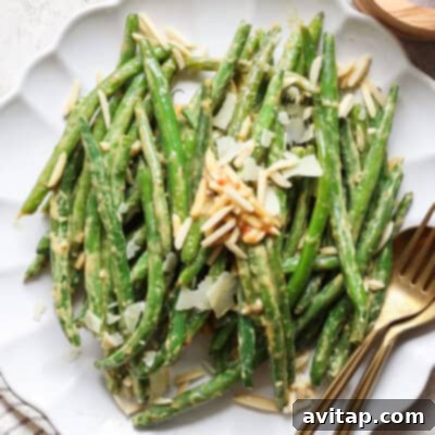 A delectable plate of vibrant green bean sauté, generously topped with golden toasted slivered almonds and a sprinkle of delicate cheese, elegantly presented with a fork and knife. This inviting dish eagerly awaits diners.