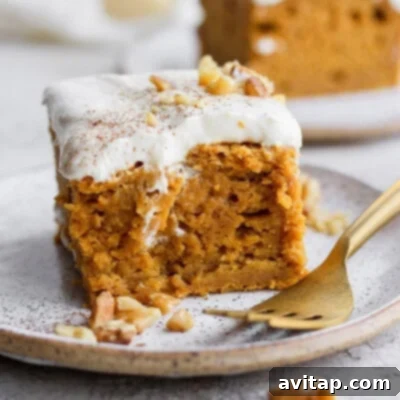A delectable slice of moist pumpkin sheet cake, generously topped with creamy white frosting and a delicate sprinkle of cinnamon. The cake is presented on a white plate, invitingly ready for dessert, promising warmth and autumnal spices.