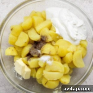 A glass bowl containing boiled, sliced potatoes, a pat of butter, a dollop of sour cream, salt, and black pepper on a light surface.