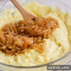 A glass bowl of mashed potatoes topped with caramelized onions, being mixed with a wooden spoon.