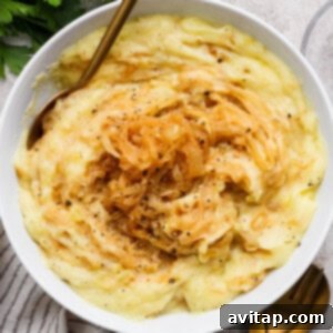 A bowl of mashed potatoes topped with caramelized onions and black pepper, with a gold fork and spoon beside the bowl on a light textured surface.
