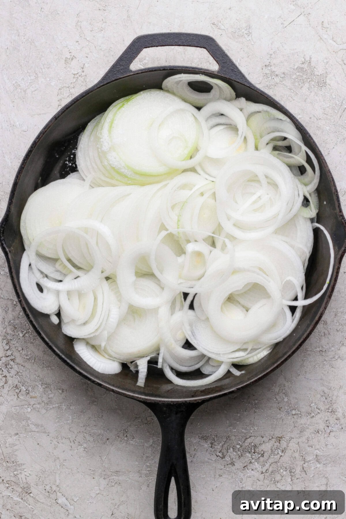 A cast iron skillet filled with raw, sliced white onions, arranged in overlapping rings on a light textured surface.