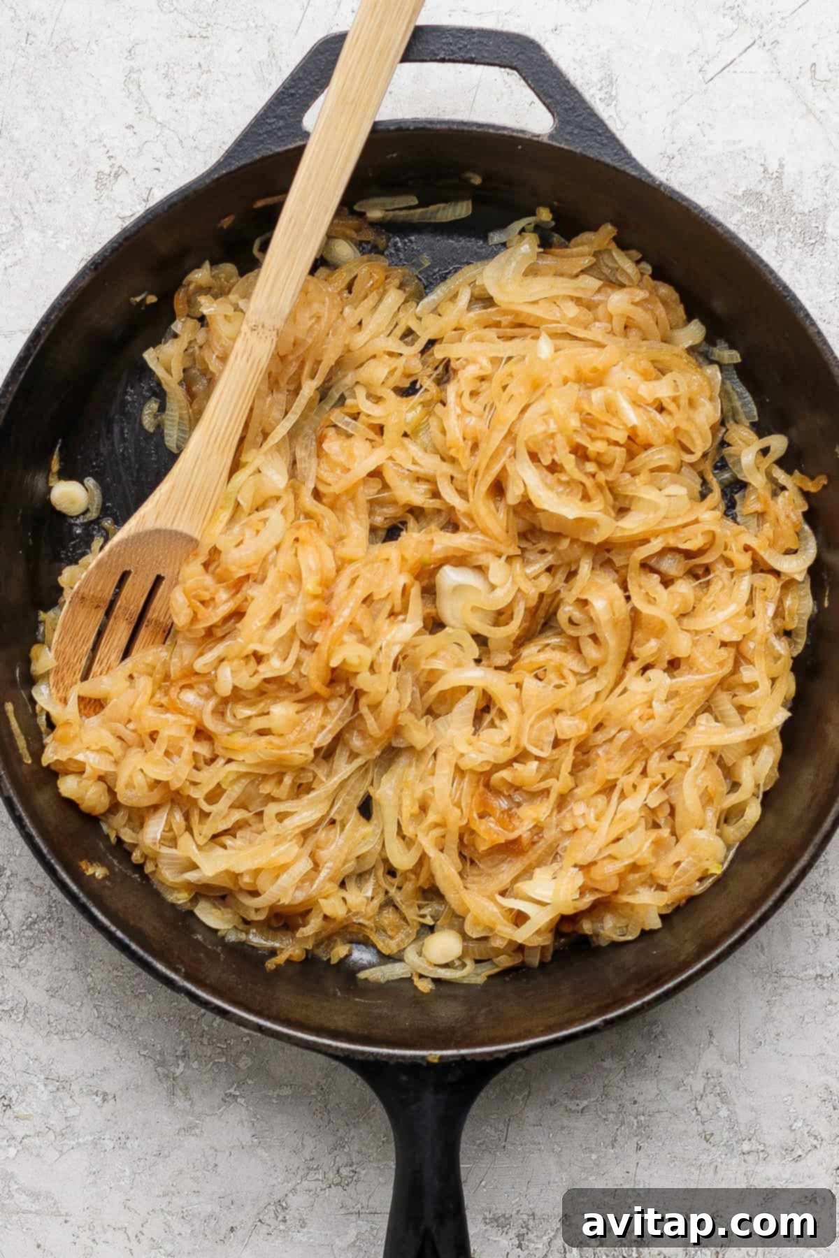 Sliced onions caramelizing in a black cast iron skillet with a wooden spoon resting on top.