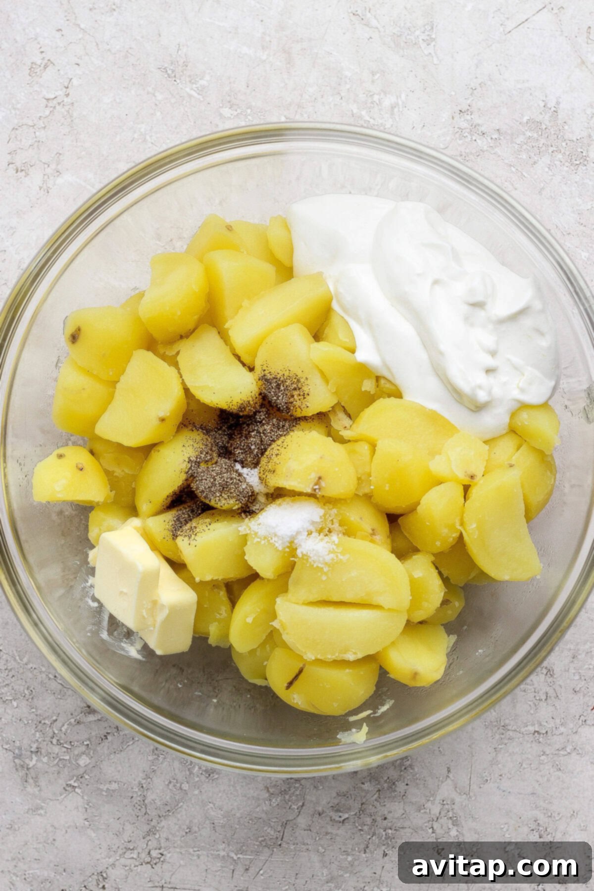A glass bowl containing boiled, sliced potatoes, a pat of butter, a dollop of sour cream, salt, and black pepper on a light surface.