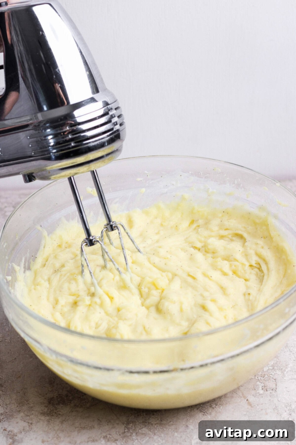 A handheld electric mixer blending creamy batter in a clear glass bowl on a light counter.