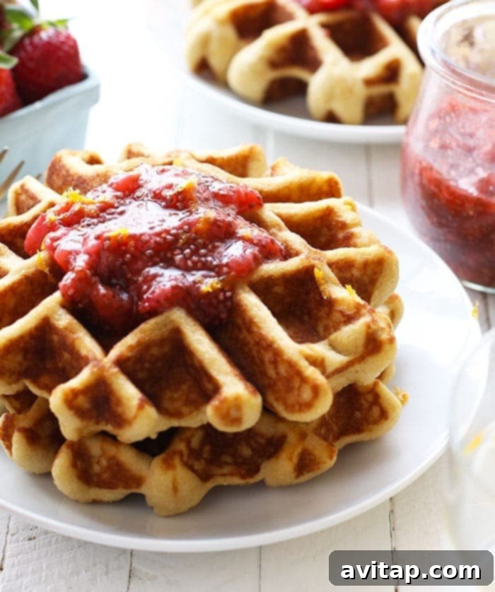 A stack of golden-brown paleo waffles, generously topped with homemade strawberry chia compote, invitingly presented on a white plate.