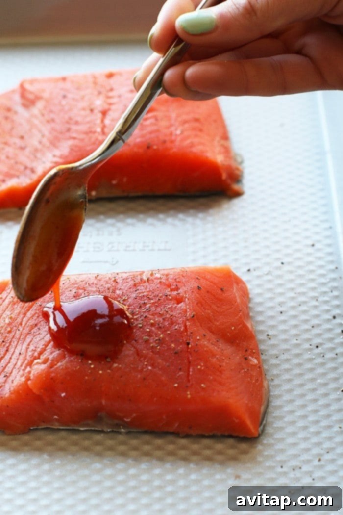 Honey Sriracha sauce being poured generously over a salmon fillet on a baking sheet, ready for broiling.