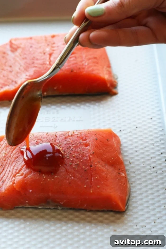 Whip up this amazing Honey Sriracha Salmon in just 8 minutes. All you need are 4 ingredients, and your oven broiler to make this protein-packed dinner that everyone will love! Pouring honey sriracha sauce over raw salmon fillets on a baking sheet, ready for broiling.