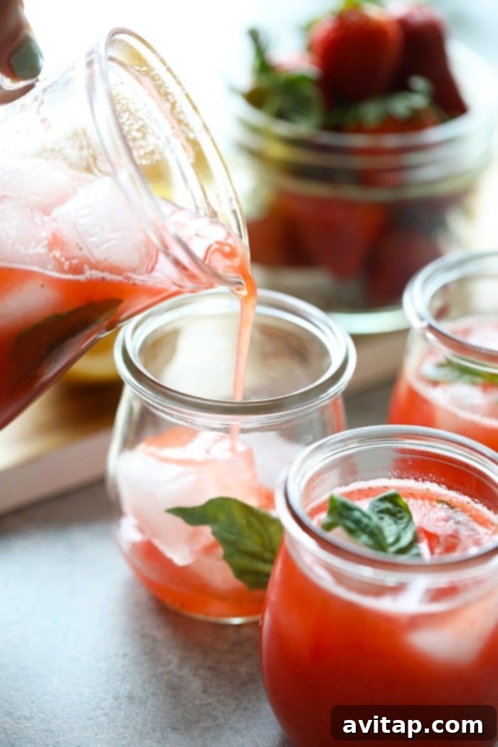 Refreshing strawberry basil lemonade being poured from a pitcher into a glass filled with ice and garnished with fresh ingredients.