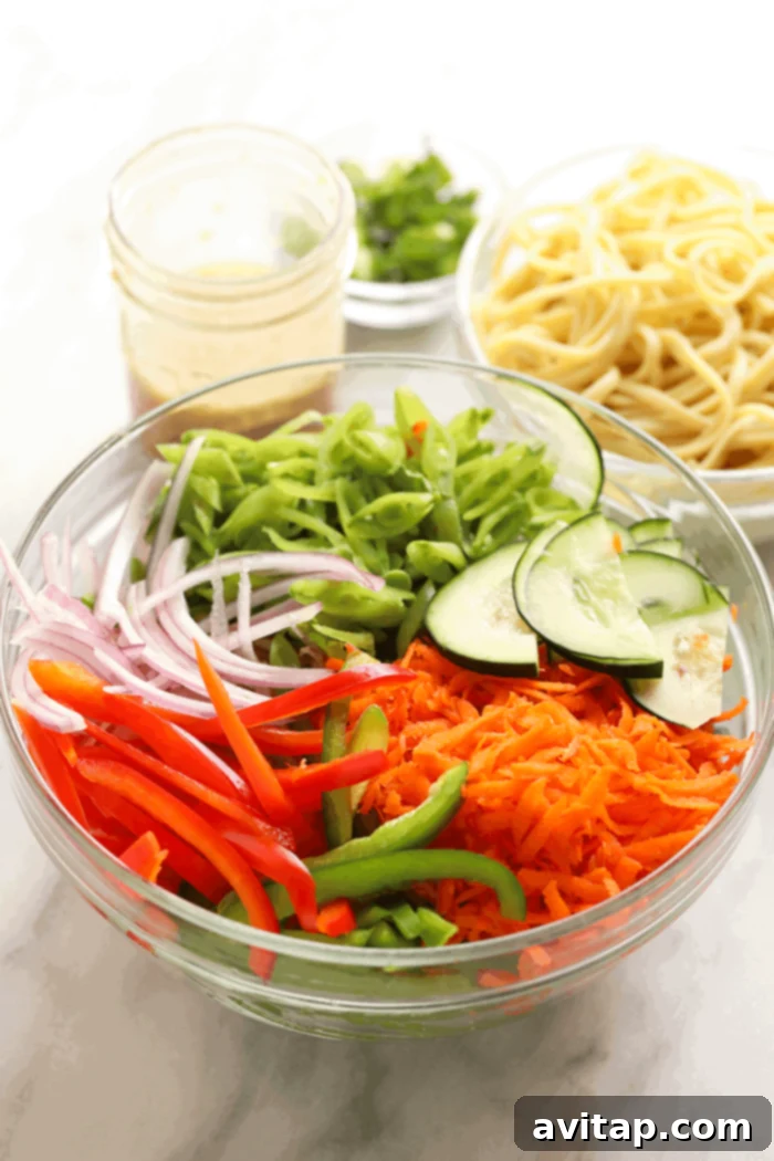 Assortment of fresh ingredients for Asian Noodle Salad, including sliced peppers, carrots, and peas, in a large mixing bowl.