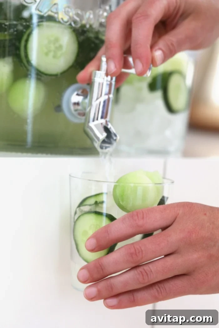 Cucumber vodka cocktails being dispensed from a large mason jar into individual glasses, garnished with fresh cucumber and mint.