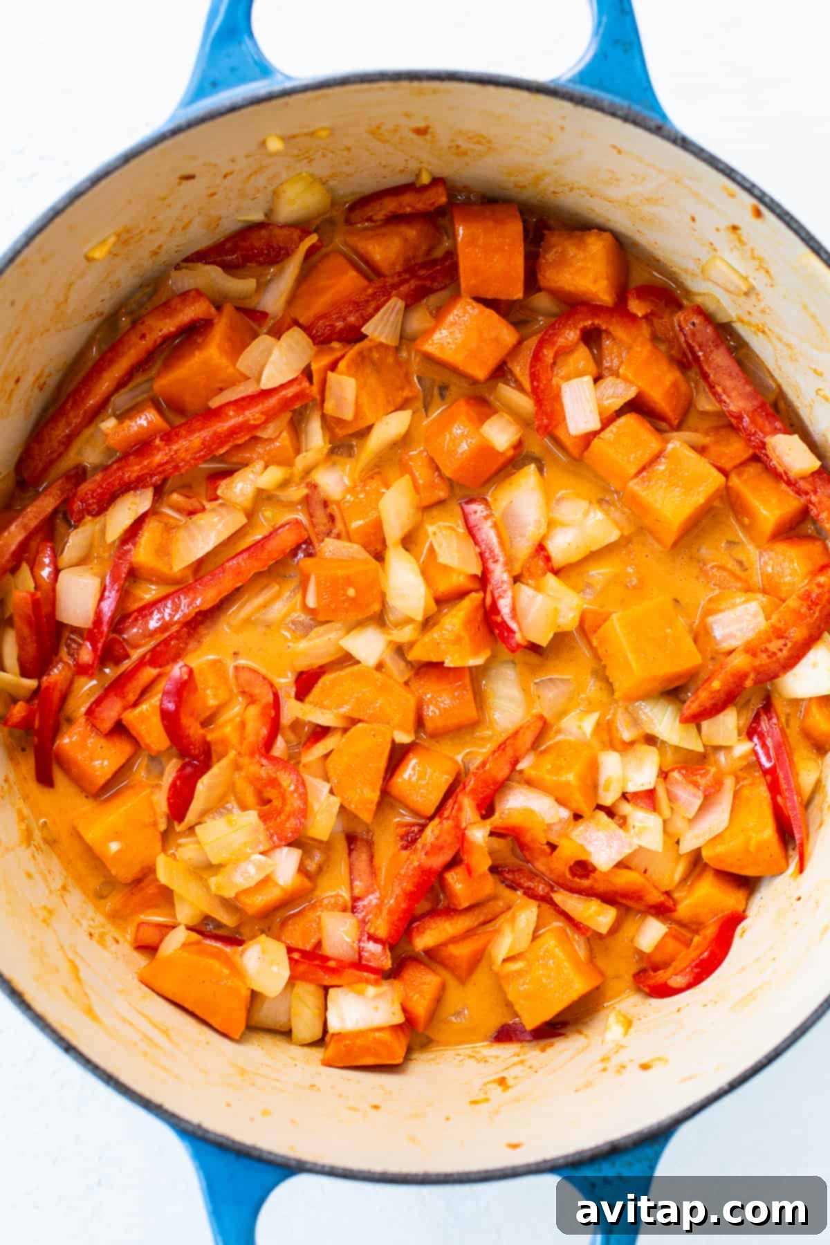 Freshly chopped sweet potatoes and colorful bell peppers, along with other vegetables, simmering in a rich, aromatic coconut curry sauce within a pot.