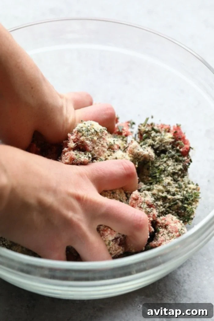 Hands mixing ground beef, breadcrumbs, herbs, and egg in a large glass bowl to prepare meatball mixture.