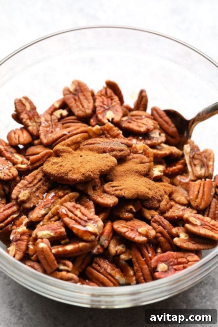 Raw pecan halves being tossed with maple syrup and ground cinnamon in a glass mixing bowl, ready for roasting.