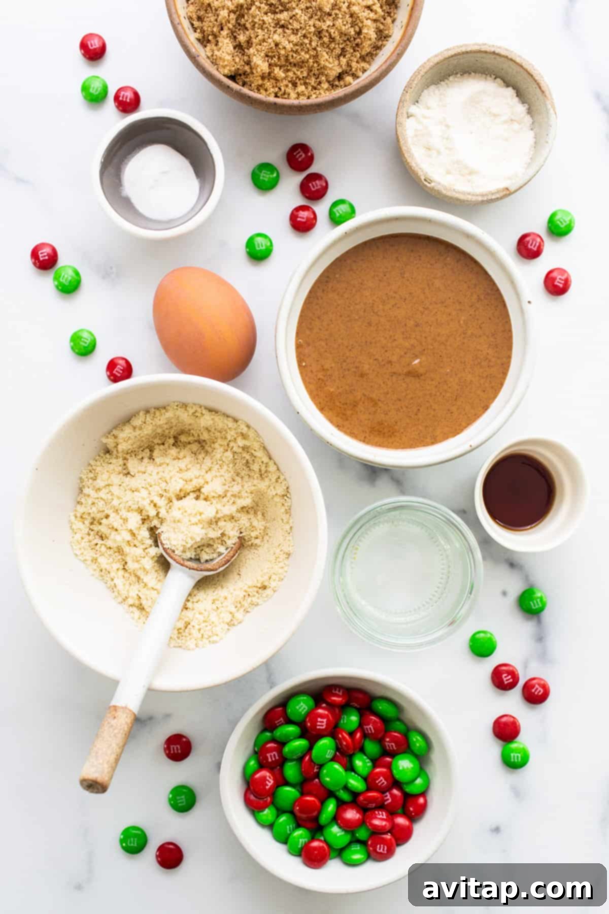 Christmas cookie ingredients laid out on a white marble table, including nut butter, flours, eggs, and M&Ms.