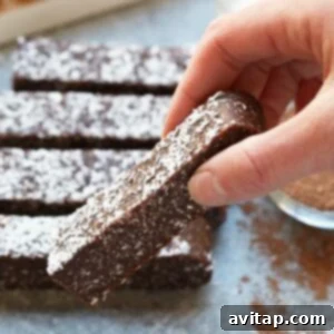 A person dipping a chocolate coconut date bar into melted chocolate, highlighting its deliciousness and potential for extra indulgence.