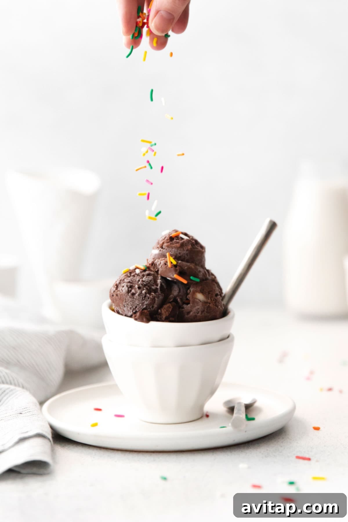 This chocolate protein ice cream recipe is dairy-free and made with frozen bananas, cocoa powder, chocolate protein powder, and almond milk! With 15 grams of protein per serving, it is a healthy treat too! Close-up of a hand gently sprinkling colorful rainbow sprinkles onto a swirl of chocolate protein ice cream, emphasizing the fun and customizable aspect of the dessert.