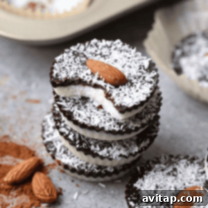 A stack of three Almond Joy Cups with a bite taken out, revealing layers of chocolate and coconut, topped with almonds. Scattered cocoa powder and almonds surround the cups.