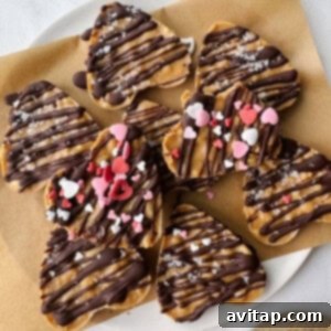Heart-shaped cookies drizzled with chocolate, sprinkled with salt, and adorned with heart-shaped sprinkles rest on a parchment-lined plate. Nestled beside them are delectable peanut butter cups, adding an irresistible touch to this sweet display.