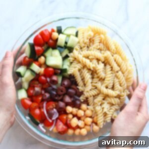 A person gently tossing a bowl of Greek pasta salad with a spoon, showing the fresh ingredients.