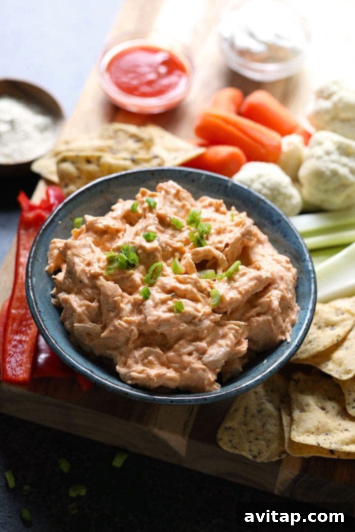 A vibrant crockpot buffalo chicken dip, garnished and ready to be served, surrounded by various dippers like celery and carrot sticks on a rustic cutting board.