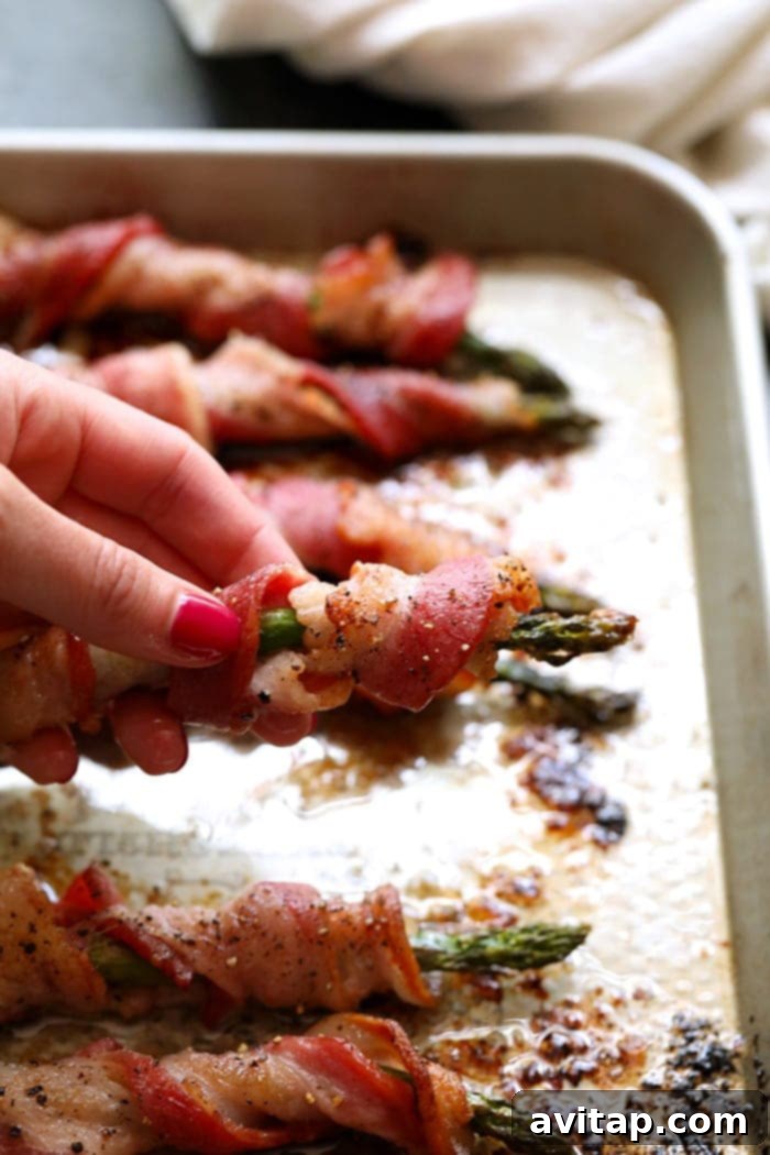 A hand presenting a single perfectly cooked bacon wrapped asparagus spear.