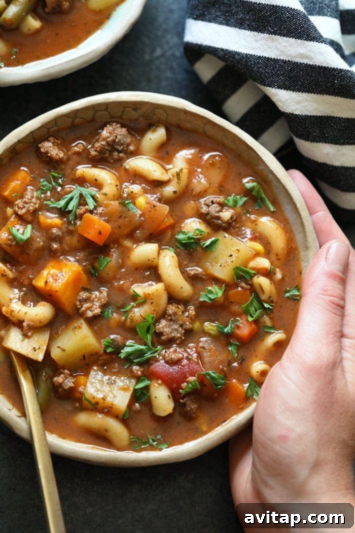 A hand holding a bowl of steaming hamburger soup, garnished with fresh herbs, showcasing a delicious and healthy meal.