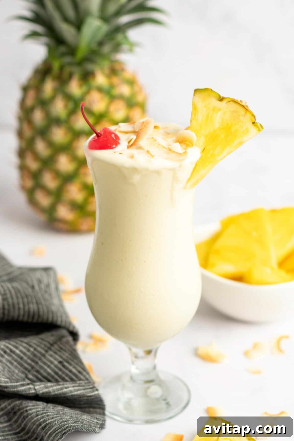 A tall glass of creamy Pina Colada smoothie garnished with a pineapple slice, a maraschino cherry, and shredded coconut, with a whole pineapple and a bowl of pineapple chunks in the background.