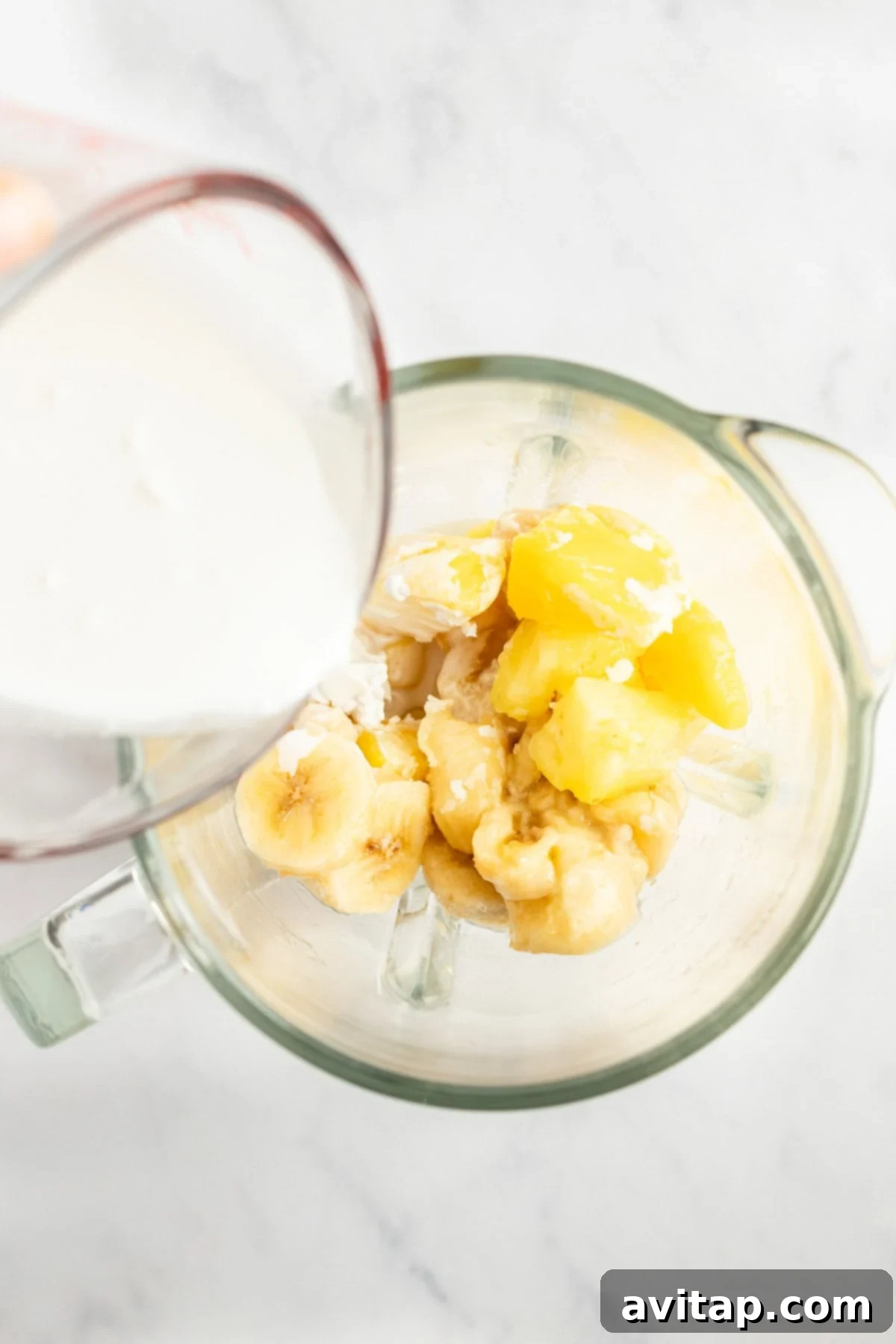 A measuring cup pours light coconut milk into a blender already containing sliced frozen bananas and chunks of frozen pineapple, ready for blending.
