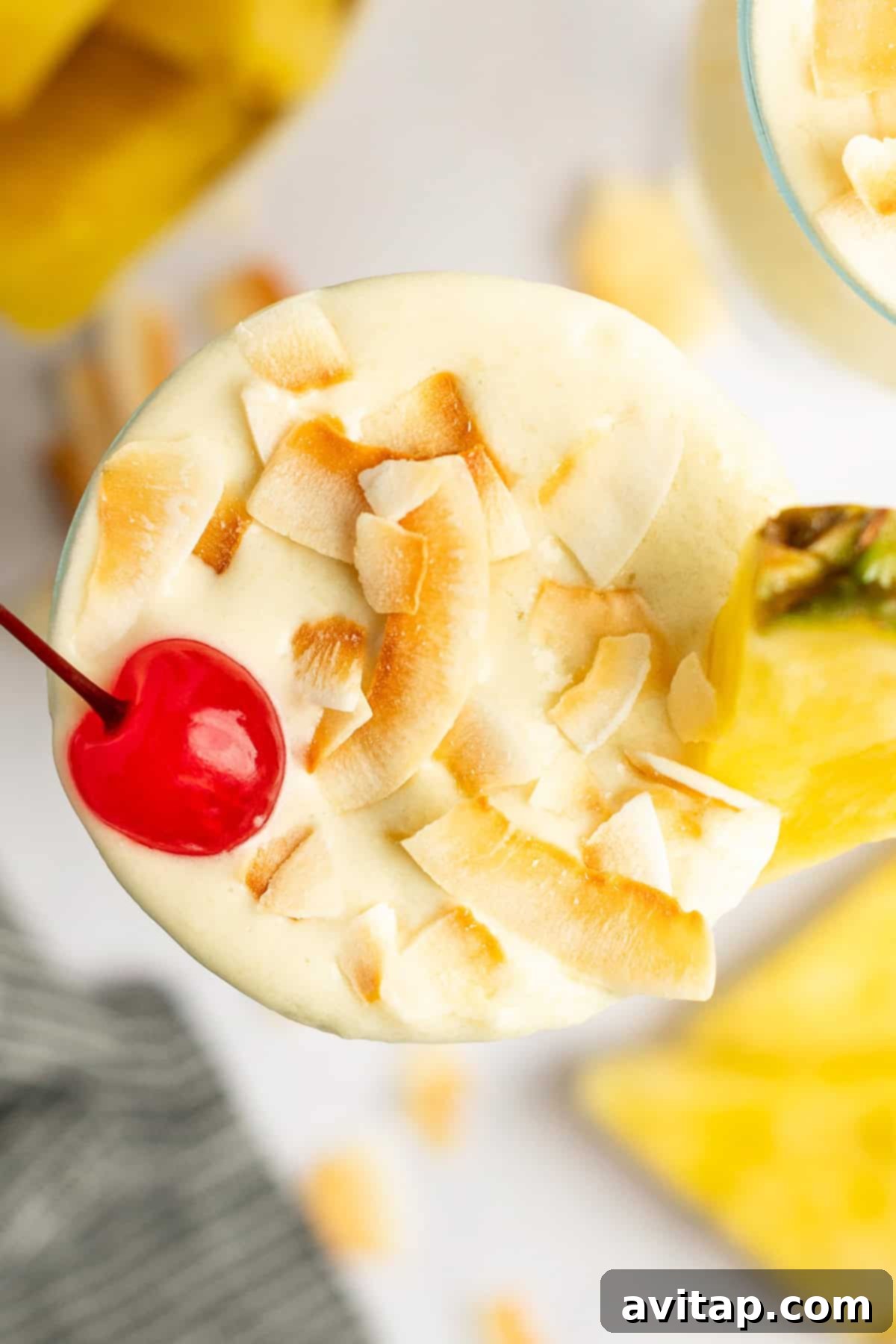 Top-down view of a creamy tropical cocktail, garnished exquisitely with golden toasted coconut flakes, a vibrant maraschino cherry, and a fresh pineapple wedge, served in a elegant glass.