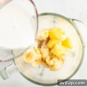A measuring cup pours milk into a blender containing sliced bananas and pineapple chunks.