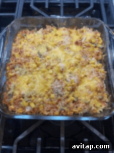 A glass baking dish brimming with a vibrant chicken casserole, featuring rice, golden corn, black beans, diced vegetables, and a luscious layer of melted cheese on top, resting on a rustic stovetop.