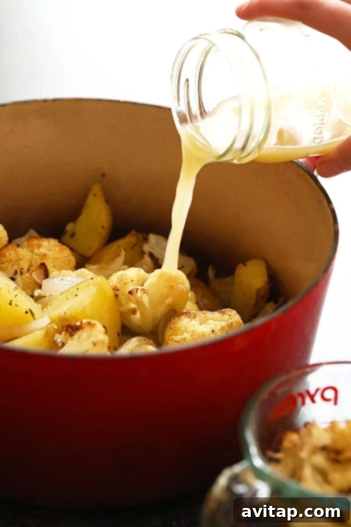 Roasted potatoes and cauliflower in a blender, with chicken broth being poured over them, ready to be blended into a creamy soup