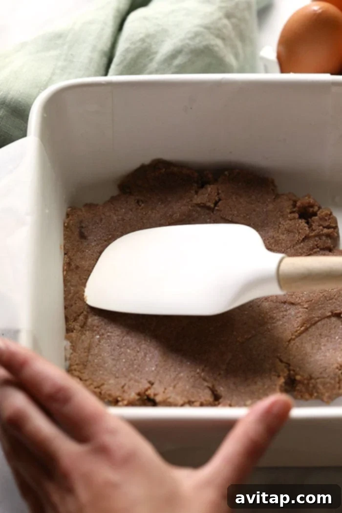 A pair of hands using a spatula to smoothly spread the prepared almond flour crust dough across the bottom of a baking pan, demonstrating the ease of making these healthy pumpkin pie bars.