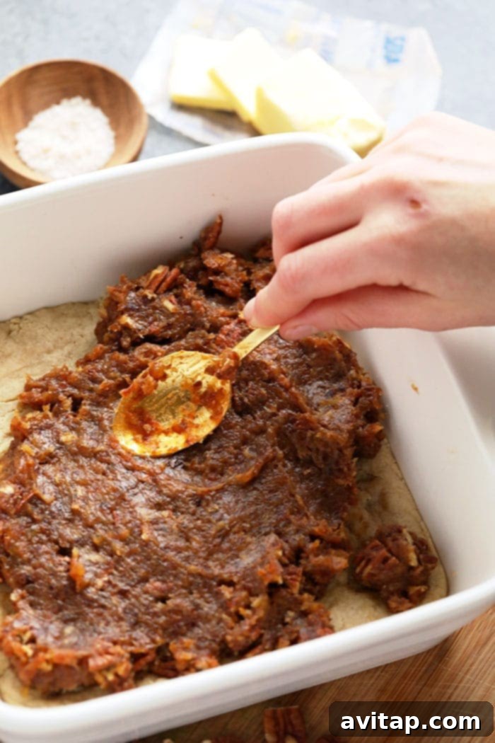 Spreading Date Caramel onto Almond Crust for Pecan Pie Bars A hand gently spreading rich date caramel evenly over a golden almond shortbread crust in a baking pan.
