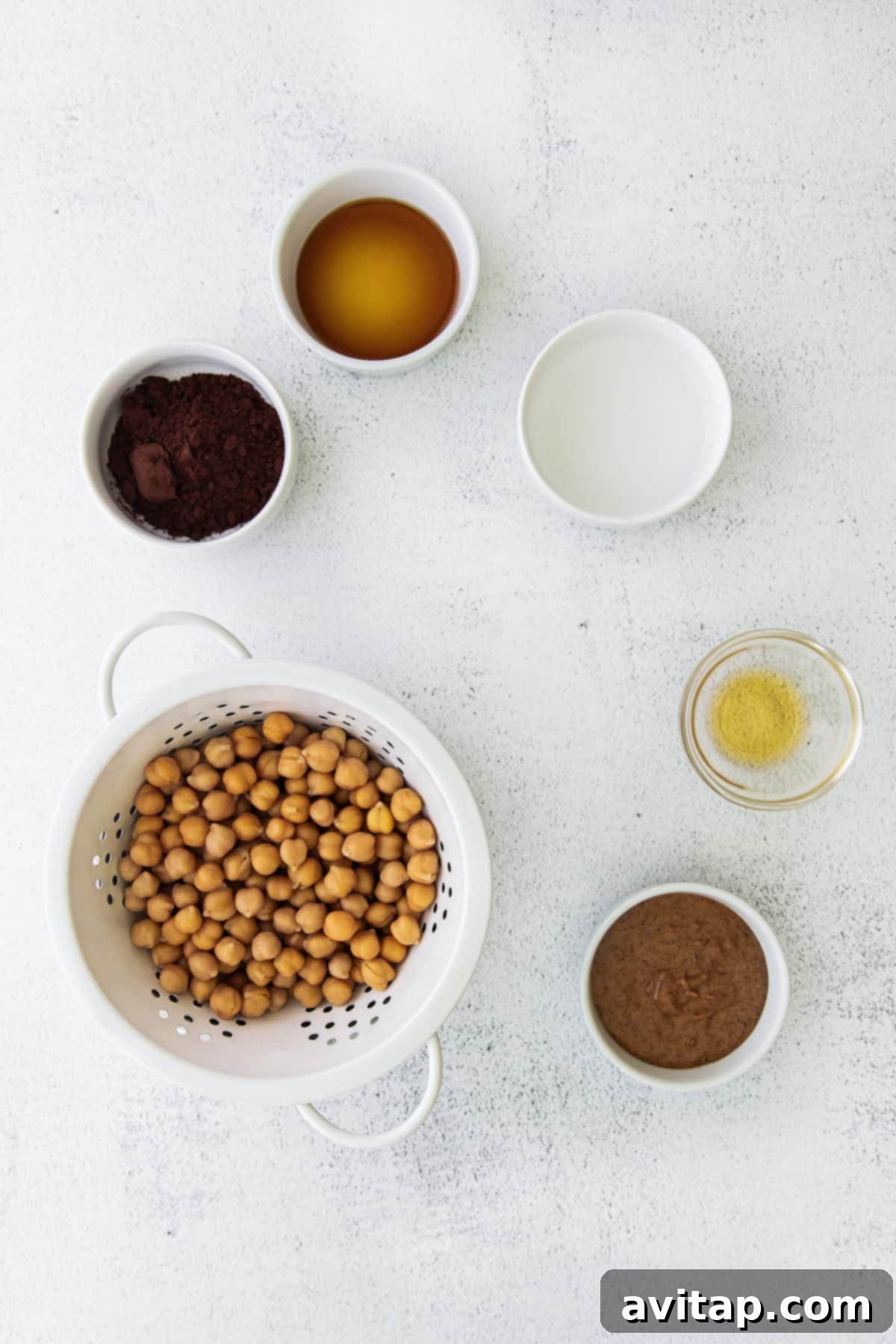 Essential ingredients for chocolate hummus: garbanzo beans, almond butter, cocoa powder, and maple syrup, neatly arranged on a kitchen countertop.