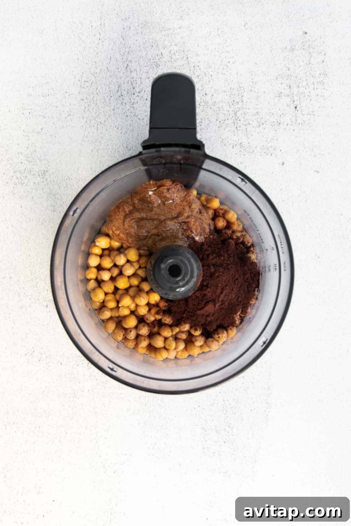 All the chocolate hummus ingredients, including chickpeas, almond butter, cocoa, and maple syrup, placed inside a high-speed food processor, ready for blending.