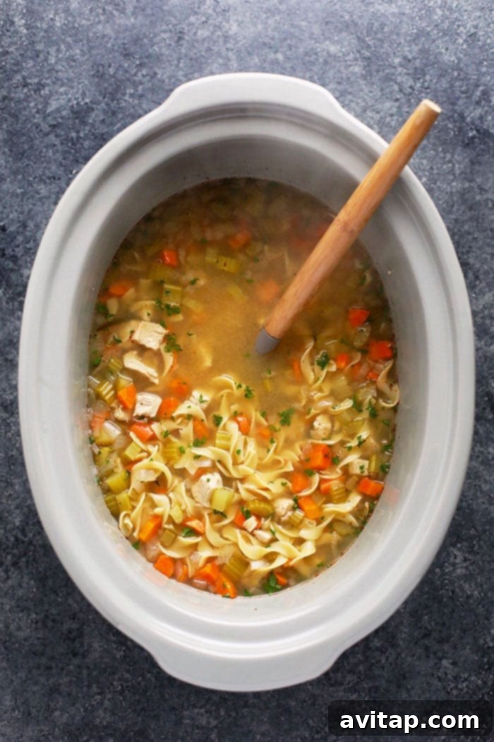 A large pot of slow cooker chicken noodle soup with a serving spoon, emitting steam and warmth.