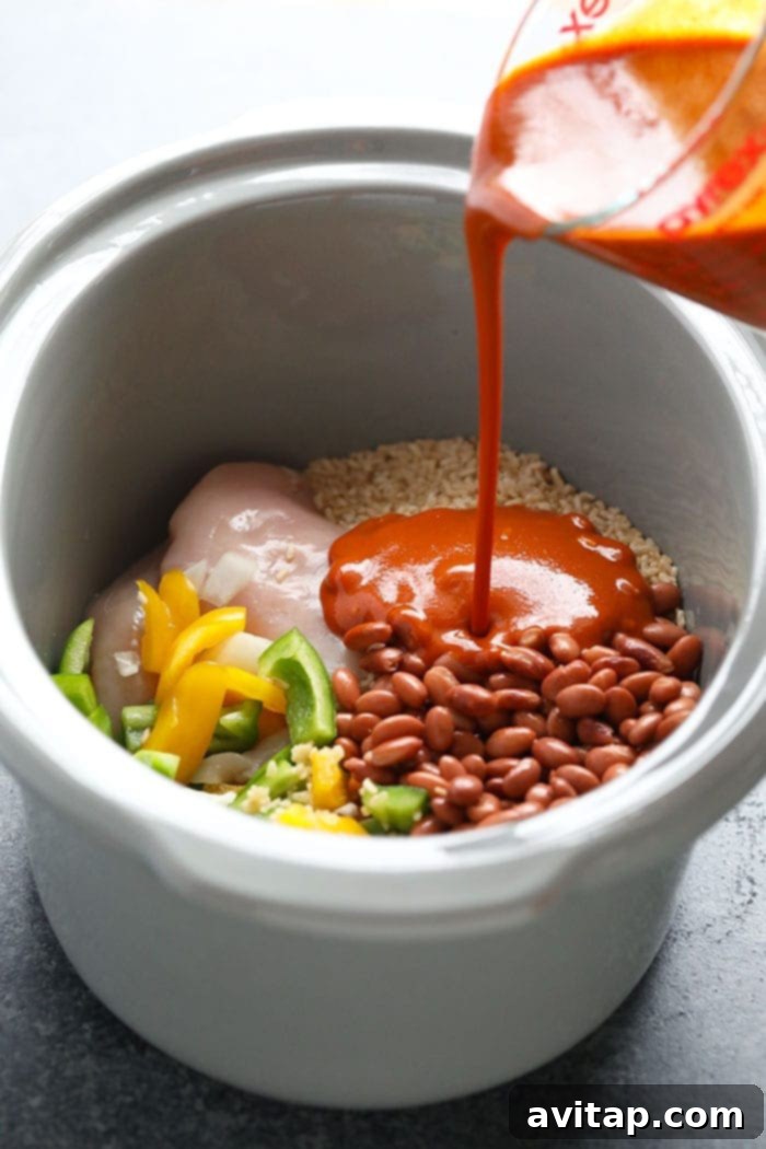 Pouring rich enchilada sauce into a slow cooker filled with chicken and vegetables.