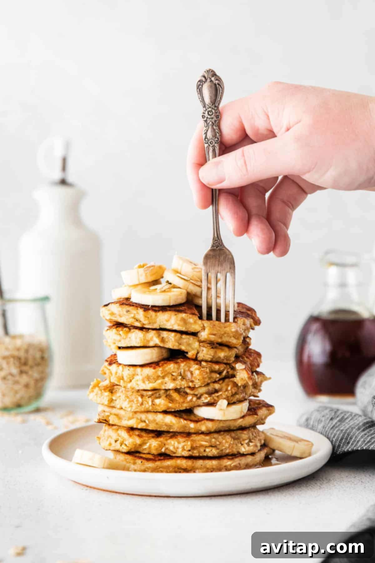 Fluffy Banana Oatmeal Pancakes: A Healthy, Gluten-Free Breakfast Favorite Fork biting into a tall stack of golden-brown banana oatmeal pancakes, drizzled with syrup and topped with sliced bananas and oats.