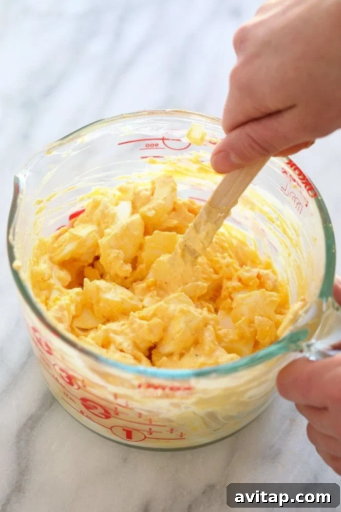 Healthy Egg Salad that packs a short list of whole ingredients and the delicious egg salad flavors you love. This healthy egg salad recipe is easy to whip up for lunches on the go, or meal prepped to enjoy all week long. Ingredients for healthy egg salad being mixed together in a large bowl. Chopped hard-boiled eggs, Greek yogurt, mayonnaise, and spices are visibly combining to create a creamy texture.