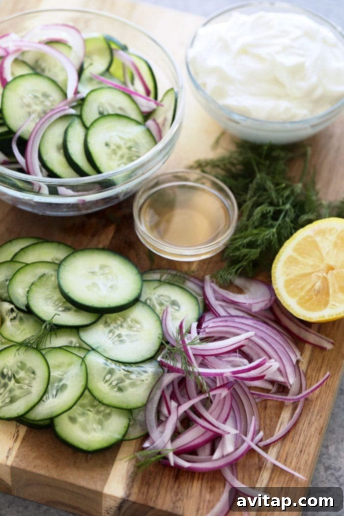 Fresh ingredients laid out on a wooden cutting board, including crisp cucumbers, red onion, a bottle of apple cider vinegar, a bowl of Greek yogurt, fresh dill sprigs, and half a lemon, ready for making the creamy cucumber salad.