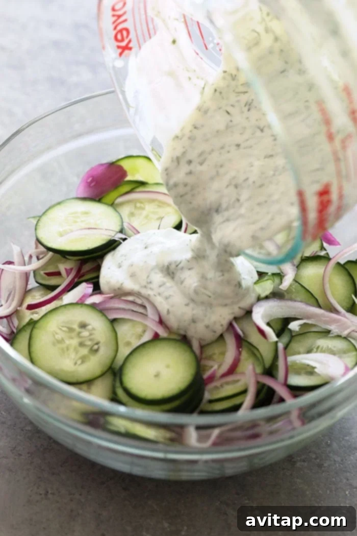 A hand gently pouring a creamy Greek yogurt dressing over a bowl of sliced cucumbers and red onion, illustrating the dressing process for the creamy cucumber salad.