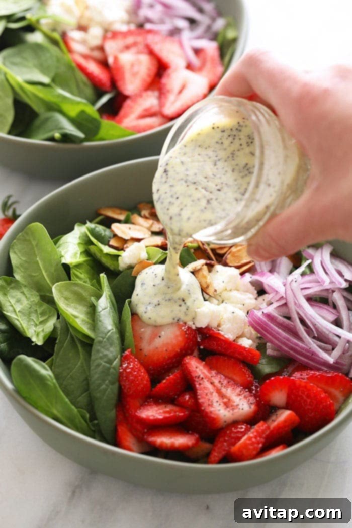 An appetizing overhead shot of rich, homemade poppy seed dressing being gracefully poured from a clear glass jar onto a vibrant Strawberry Spinach Salad, enhancing its appeal.