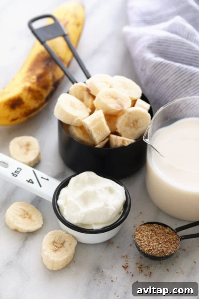 All the core ingredients for a healthy banana smoothie laid out: perfectly frozen banana slices, a container of plain Greek yogurt, a small bowl of ground flax seeds, and a glass of unsweetened almond milk.