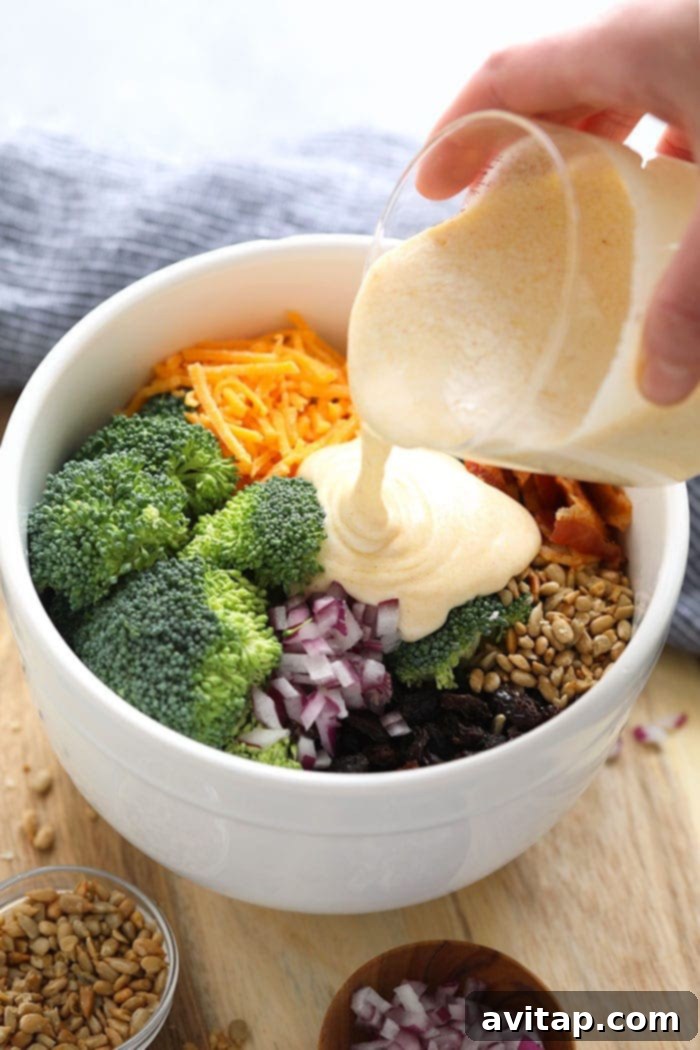 Pouring the creamy, no-mayo dressing over the fresh broccoli salad ingredients in a large bowl.