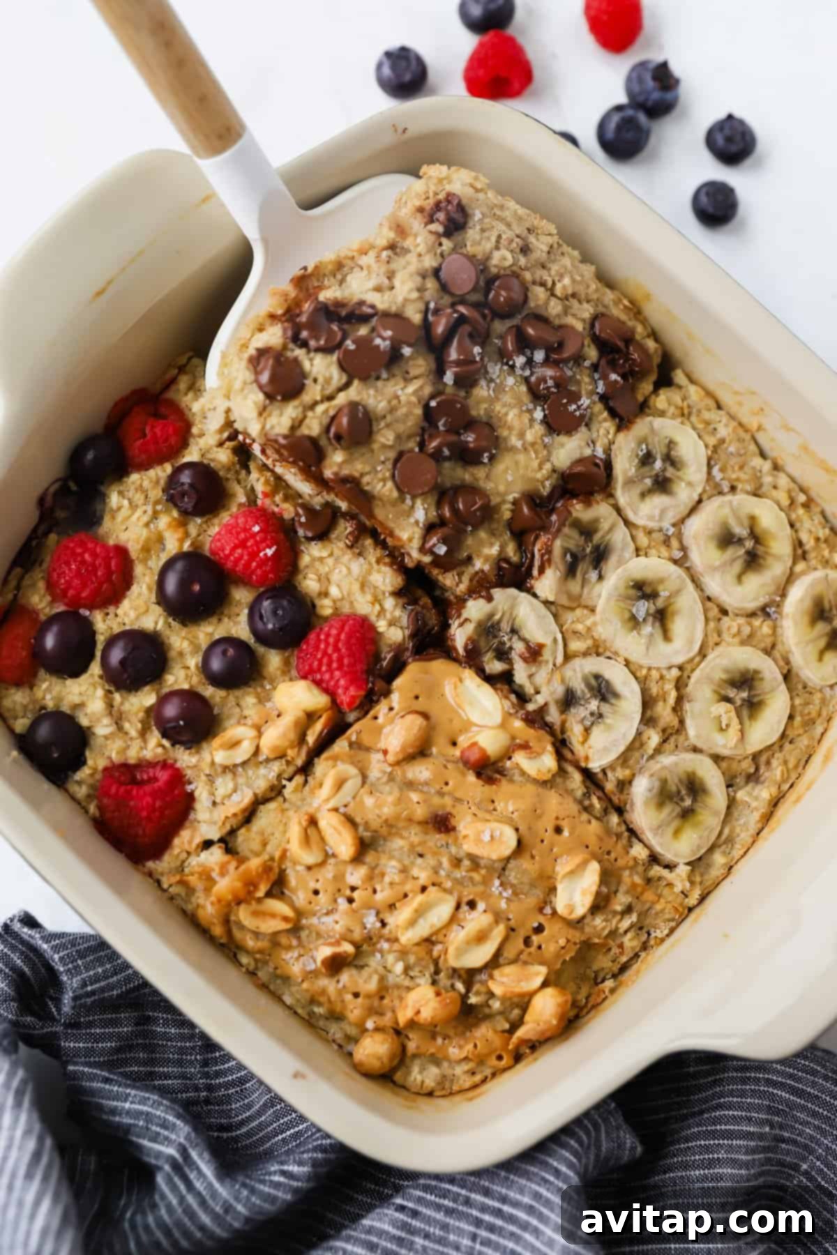 A baking dish with four sections of baked oatmeal topped with chocolate chips, banana slices, mixed berries, and peanut butter with peanuts. A spatula rests on one section, inviting a taste.